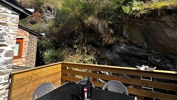 Un terrazzo in legno con tavolo e sedie, con vista su un pendio roccioso e vegetazione.