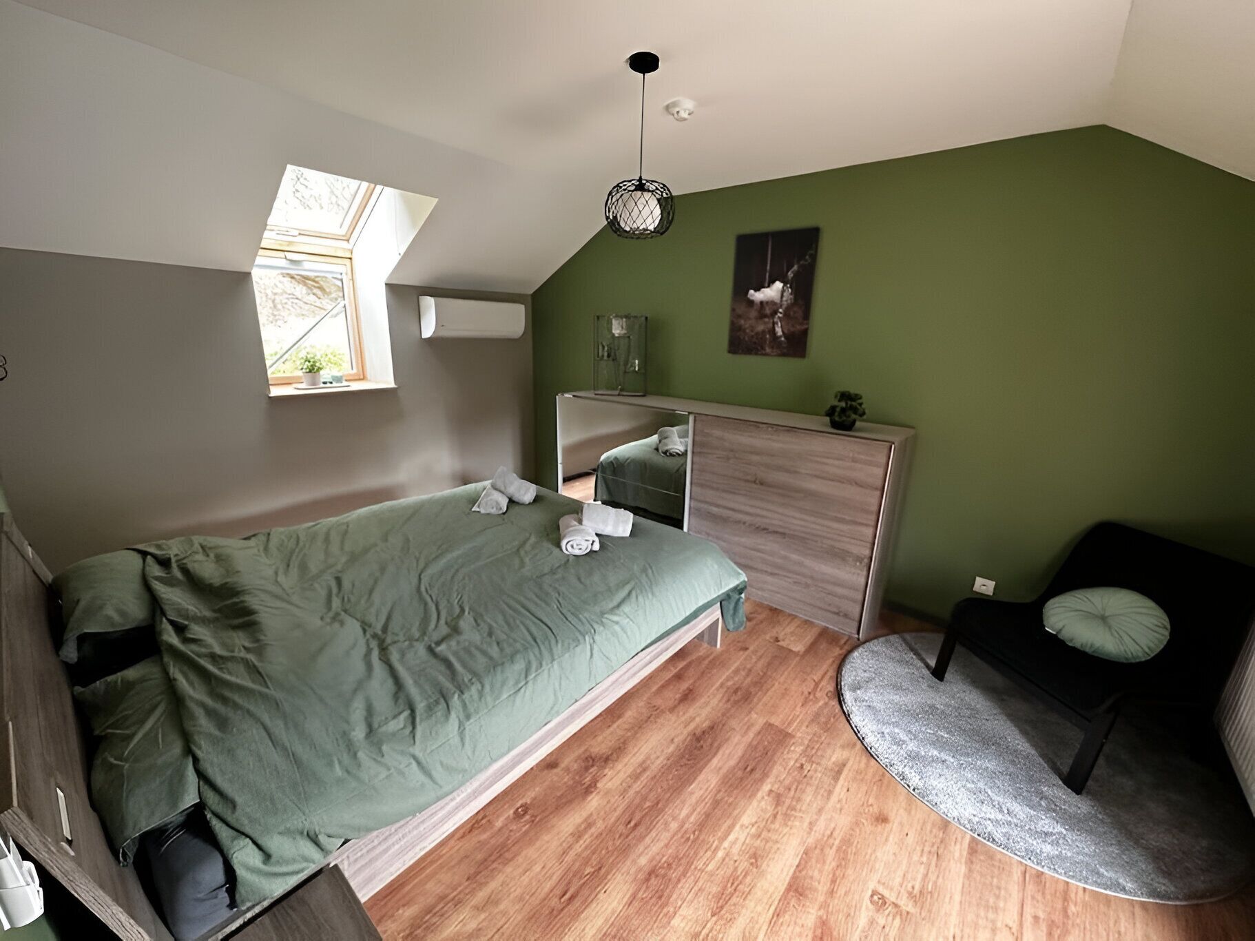 A compact bedroom with a bed, a small seating area, a window, and a green wall.