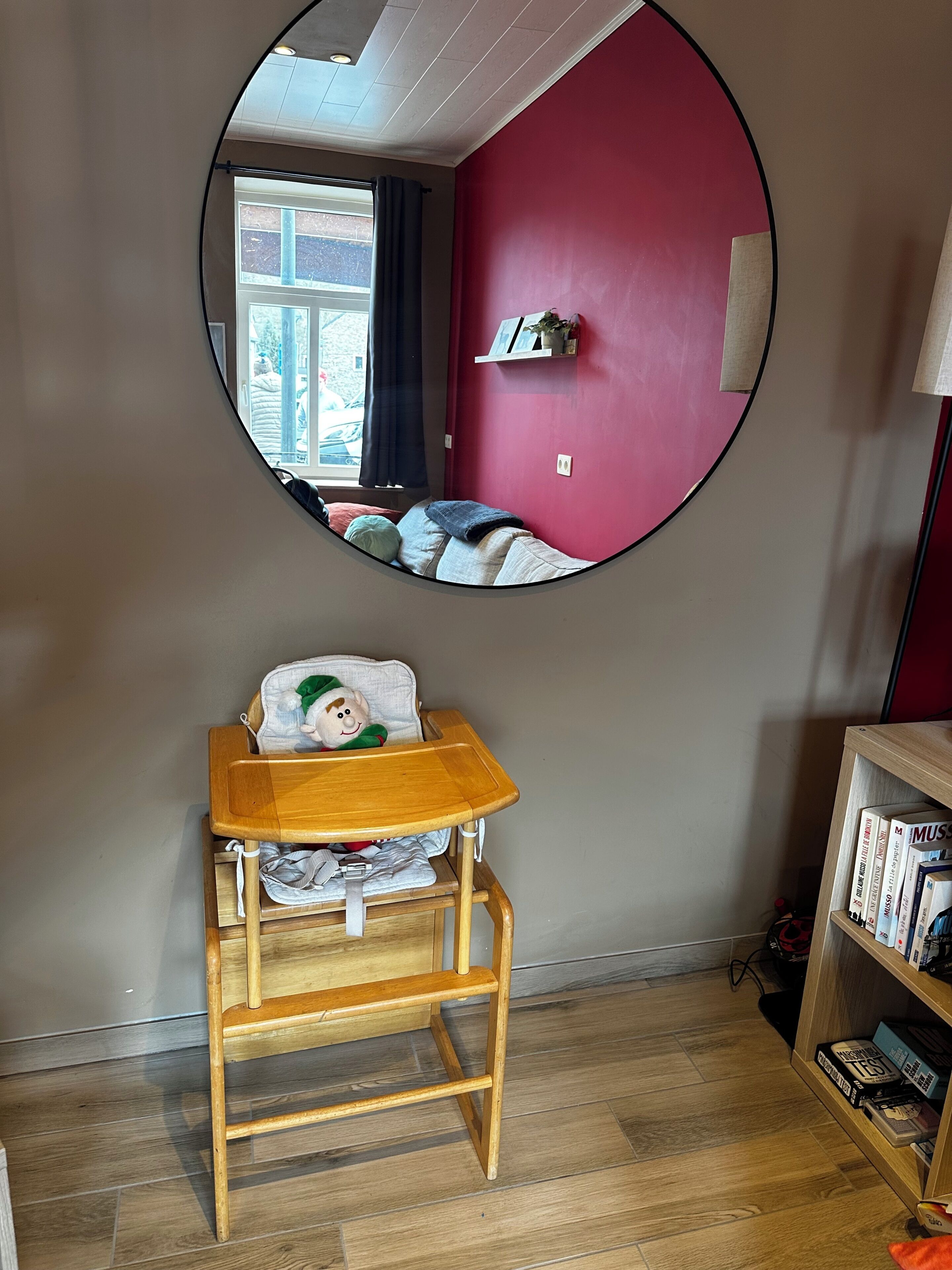 A room with a circular mirror, a wooden chair, a bookshelf, and a window with curtains.