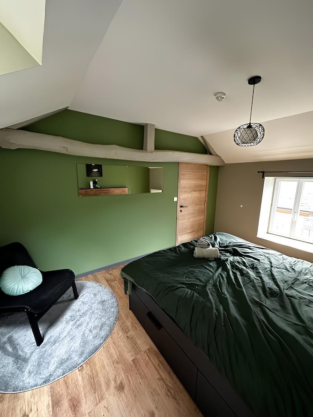 Une chambre avec un lit, une chaise, un tapis rond, un plancher en bois, un mur vert, une porte en bois, un luminaire suspendu et une fenêtre.