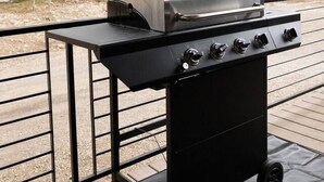 A portable grill with four burners and a lid, placed on a black metal table with wheels, on a covered patio with a metal railing. - NEW Mountain Modern Cabin (Unit 4) (Island Park)