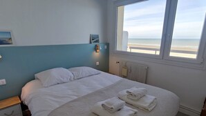 Een slaapkamer met een groot bed, twee nachtkastjes, een raam met uitzicht op het strand en een radiator.