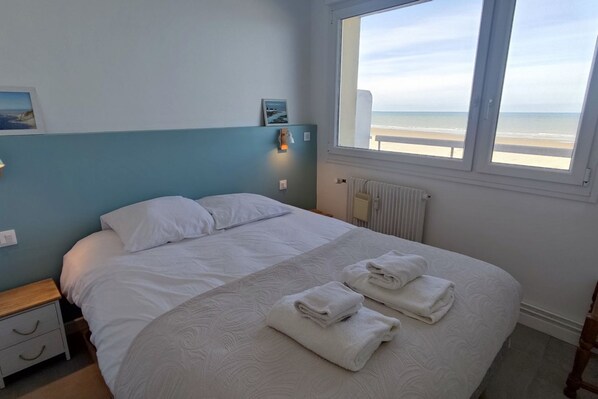 Een slaapkamer met een groot bed, twee nachtkastjes, een raam met uitzicht op het strand en een radiator.