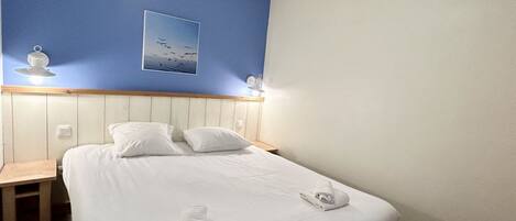 A neatly made bed with white linens and towels, a wooden nightstand, a blue wall with a framed picture, and a white ceiling.