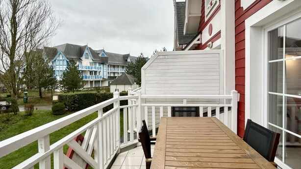 A wooden deck with a white railing, a table, and chairs. In the background, there are houses with blue and white exteriors.