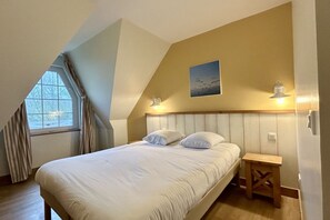 Une chambre avec un plafond en pente, un lit recouvert de linge de lit blanc, une petite table de chevet en bois et une fenêtre avec des rideaux.