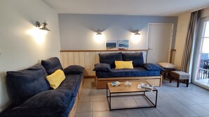 Ett vardagsrum med två mörkblå soffor, ett träbord och en liten träbänk. På väggen sitter infällda lampor och i bakgrunden syns en dörr.