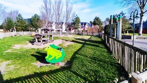 A playground with a green frog play structure, a wooden fence, and houses in the background. - Appartement Belle Dune 2 Chambres (Fort-Mahon-Plage)