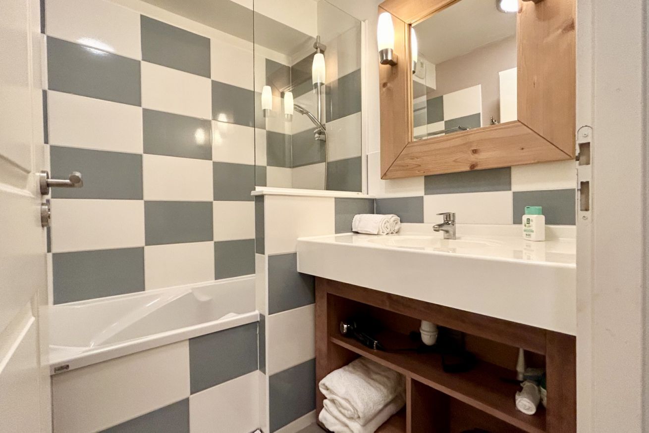 Un baño con pared de baldosas a cuadros, un lavamanos blanco con tocador de madera y ducha con cerramiento de vidrio.