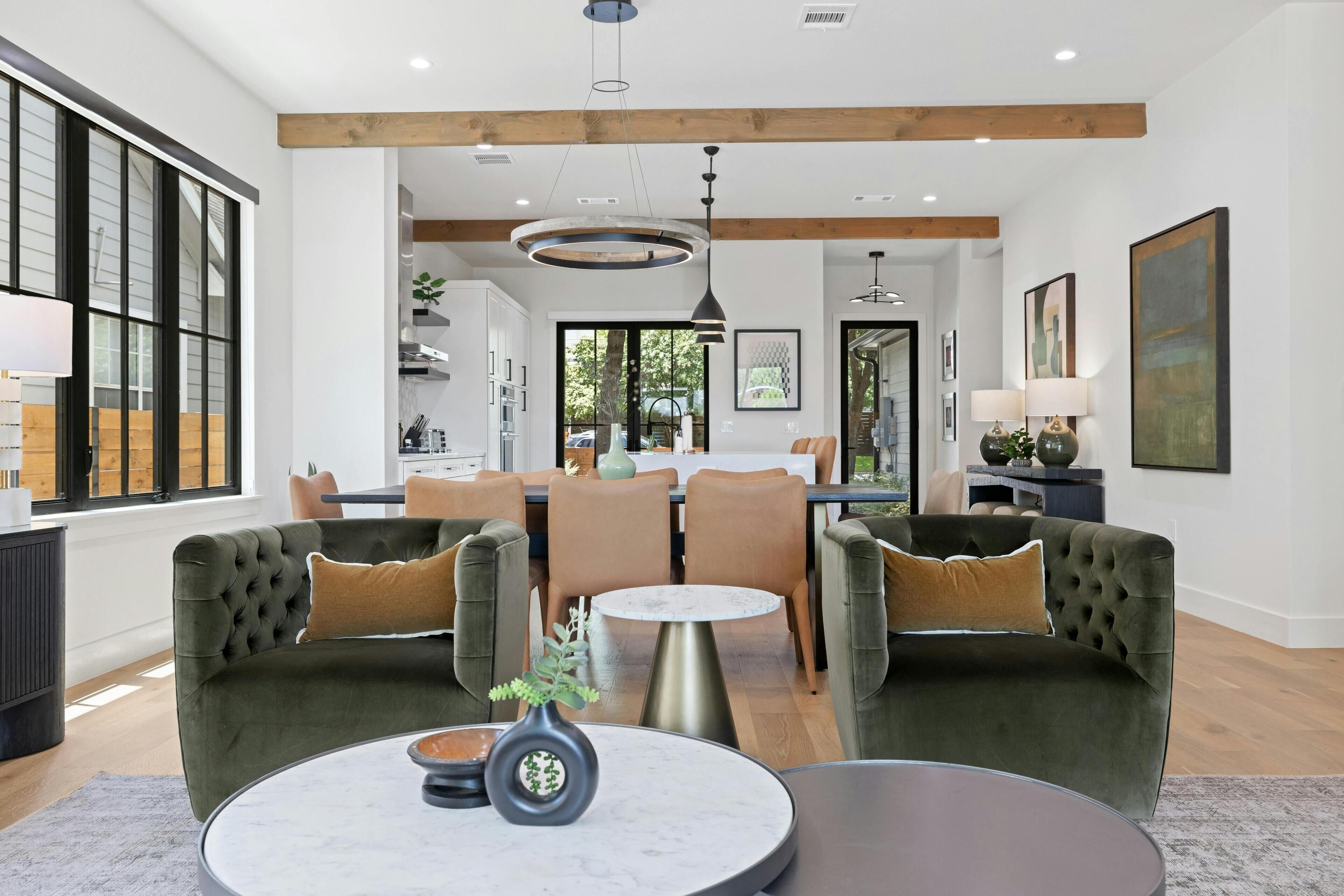 A spacious living room with a large window, a dining area with a round table, and a kitchen in the background.