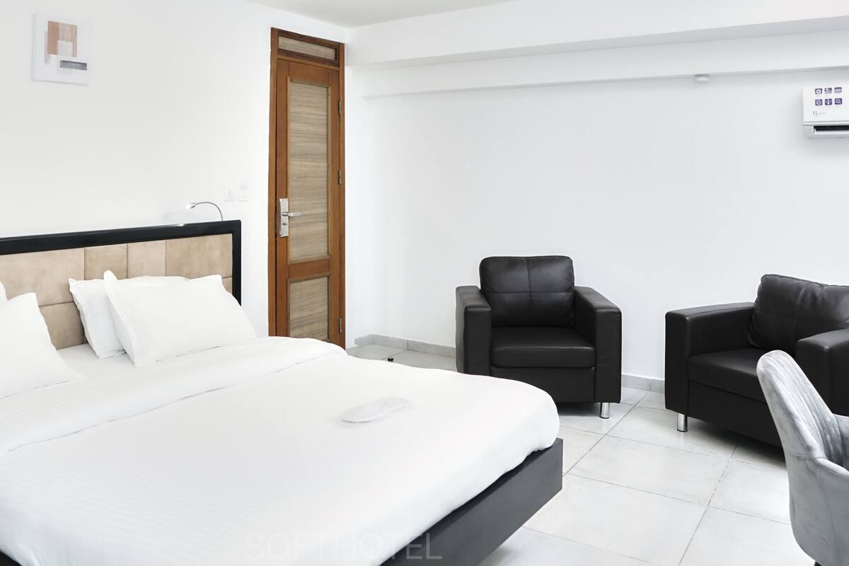 A modern hotel room with a large bed, two black leather chairs, a wooden door, and a wall-mounted air conditioning unit.