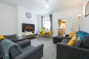 Ein modernes Wohnzimmer mit Fernseher, grauen Sofas, einem Essbereich mit gelben Stühlen und einer durch eine offene Tür sichtbaren Küche.