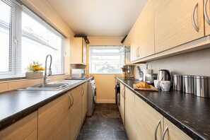Une cuisine compacte avec des meubles en bois, un plan de travail noir, un lave-linge encastré et une fenêtre avec des stores.