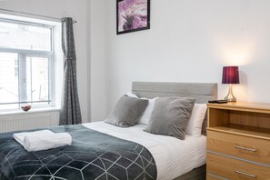 Un letto ben rifatto con una coperta dal motivo geometrico, una lampada da comodino e un tavolino di legno.