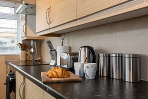 Une cuisine avec des meubles en bois, un plan de travail noir, une planche à découper avec un croissant, une bouilloire, un grille-pain et des mugs en acier inoxydable.