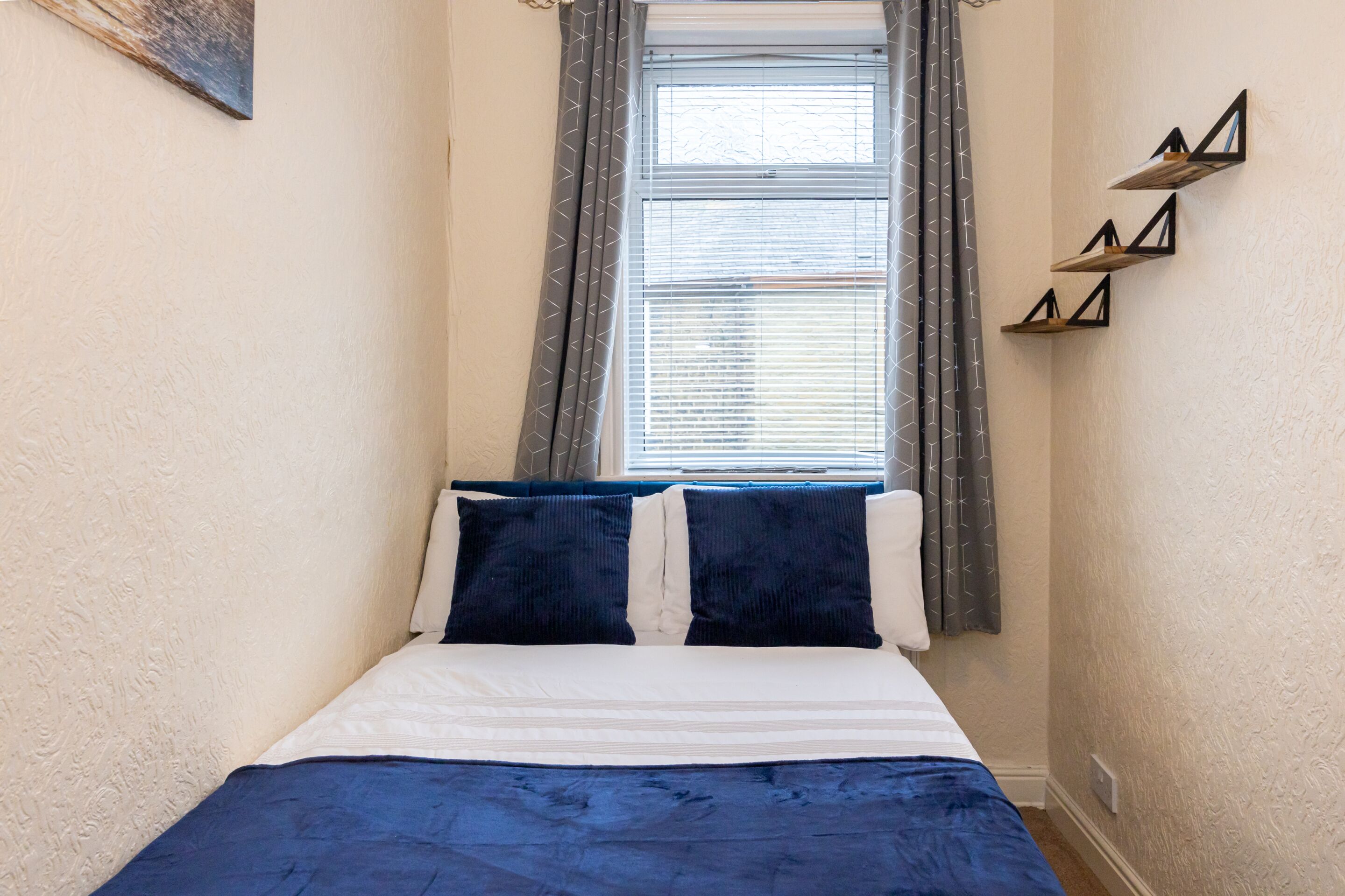 A bedroom with a bed, two pillows, a window with blinds, and a wall-mounted shelf.