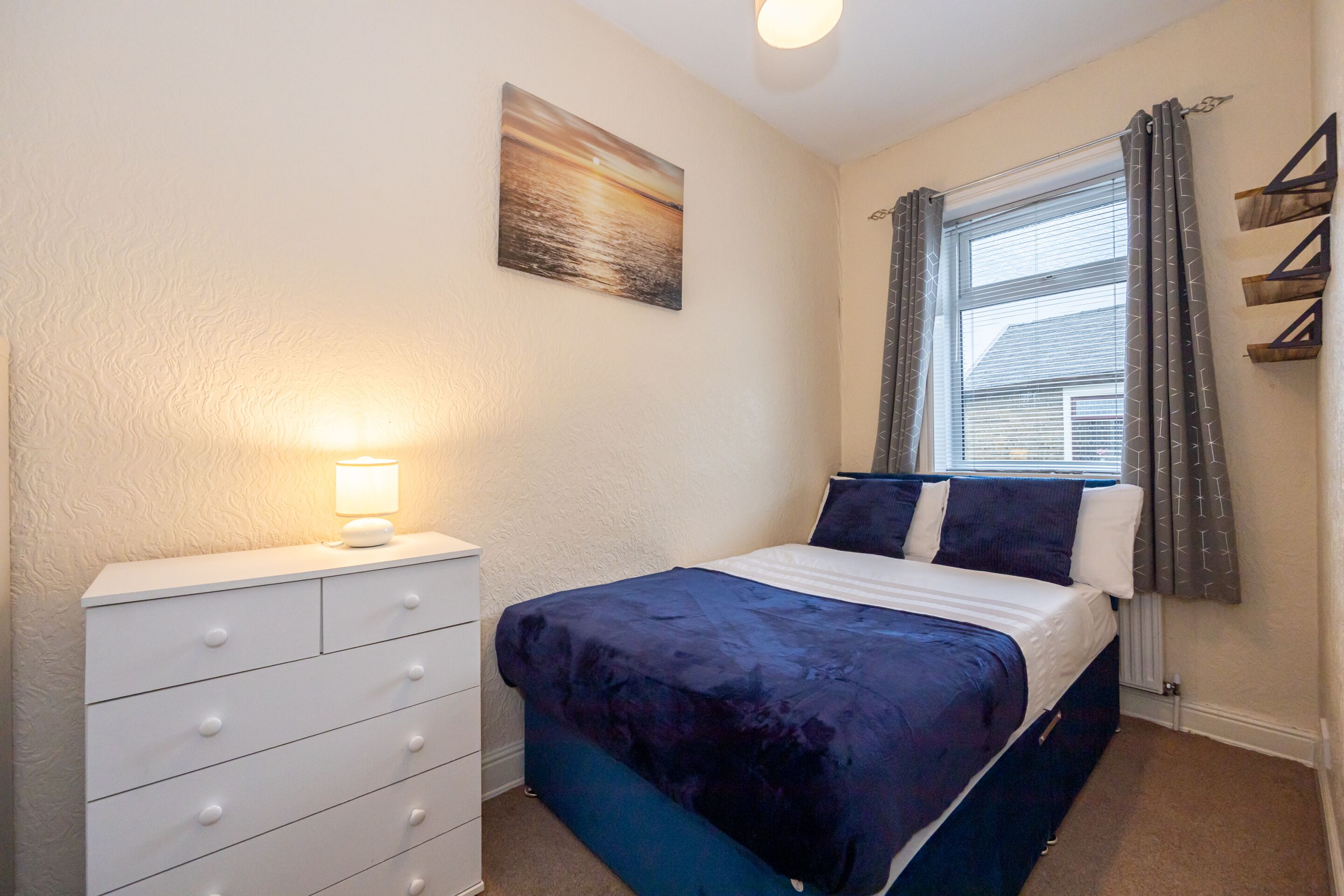 Un dormitorio con una cama, un tocador, una ventana con cortinas y un cuadro en la pared.