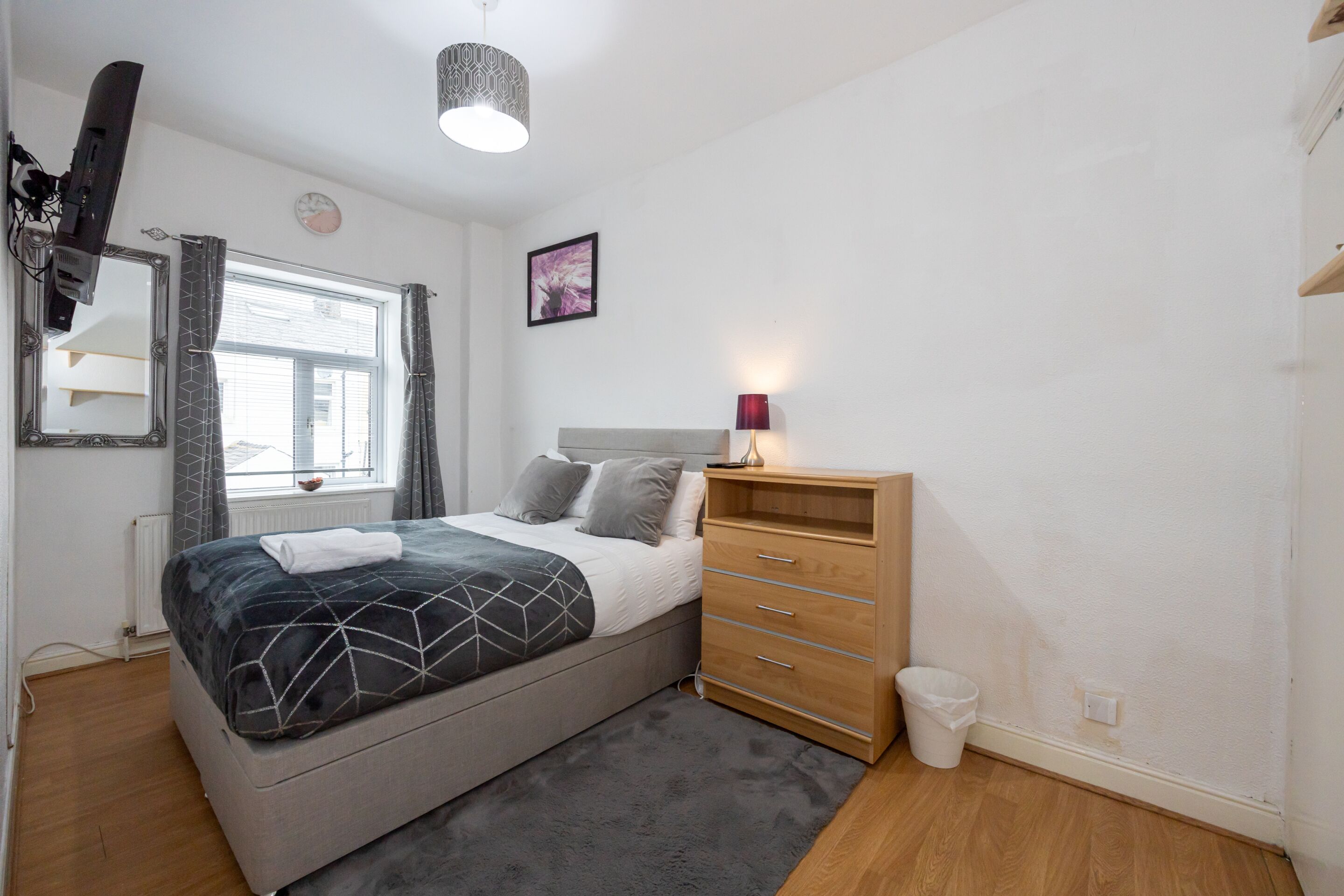 Une chambre à coucher comprenant un lit, une table de chevet avec une lampe, une commode, une fenêtre avec des rideaux et une horloge murale.