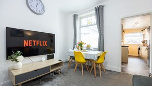 Un salon moderne doté d’un téléviseur à écran plat, d’un coin repas avec des chaises jaunes et d’une cuisine visible à travers une porte ouverte.