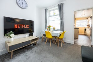 Moderni olohuone, jossa on litteänäyttöinen televisio, ruokailutila, jossa on keltaiset tuolit, ja avoimen oven kautta näkyvä keittiö.