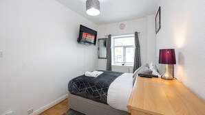 Une chambre à coucher avec un lit, un téléviseur fixé au mur, une table de chevet avec une lampe et une fenêtre avec des rideaux.