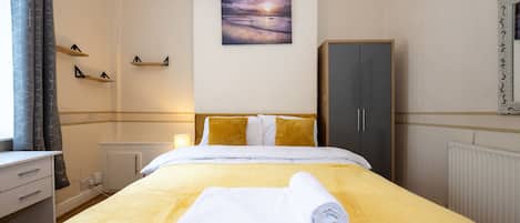 A hotel room with a bed, a painting of a sunset, a wardrobe, and a desk with a lamp.