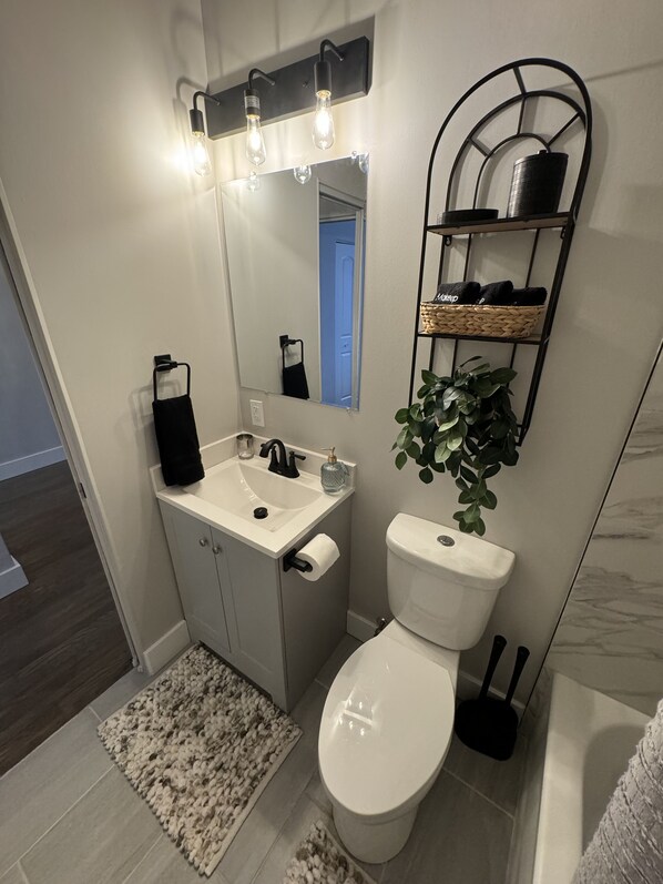 A modern bathroom with a white sink, mirror, and toilet. There is a black towel rack, a plant, and a shelf with toiletries. - Miller Beach Family Getaway (Gary)
