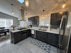 A modern kitchen with dark cabinets, a stainless steel refrigerator, a gas stove, and pendant lighting. - Miller Beach Family Getaway (Gary)