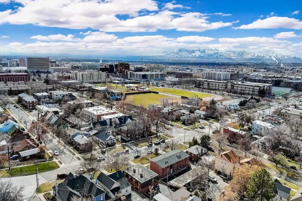 Vista aérea de una ciudad con edificios residenciales y comerciales, un extenso campo abierto y montañas nevadas al fondo.