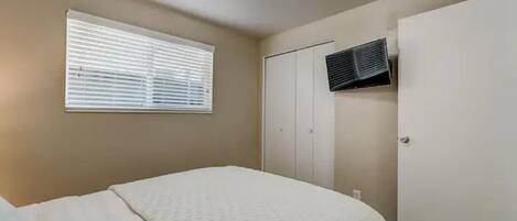 A bedroom with a bed, a window with blinds, a wardrobe, and an air conditioning unit.