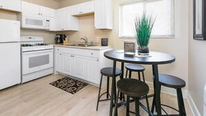 A compact kitchen with white cabinets, a stove, a microwave, and a sink. There is a small round table with stools and a potted plant on the windowsill. - Lovely top floor corner apt in trendy SugarHouse (Salt Lake City)