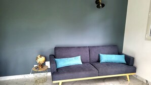 Una habitación con un sofá azul oscuro, dos cojines azul claro, una mesita redonda con base de hilo dorado y una lámpara fijada a la pared.
