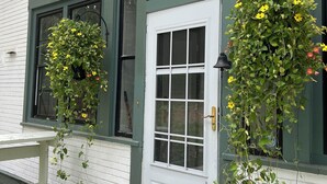 Un bâtiment blanc orné de bordures vertes, avec une porte blanche et deux fenêtres décorées de jardinières suspendues.