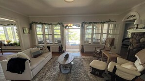 Un salon spacieux avec un canapé blanc, une table basse et une cheminée. De grandes fenêtres laissent entrer la lumière naturelle et offrent une vue sur l’extérieur.