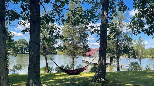 A hammock between trees by a lake with a dock and a building in the background. - Unplug and unwind at your Leonardtown waterfront vacation home! (Leonardtown)