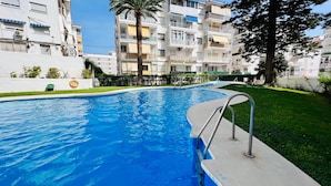 Una piscina con escalera metálica, rodeada de un césped cuidado y un edificio residencial de varios pisos al fondo.