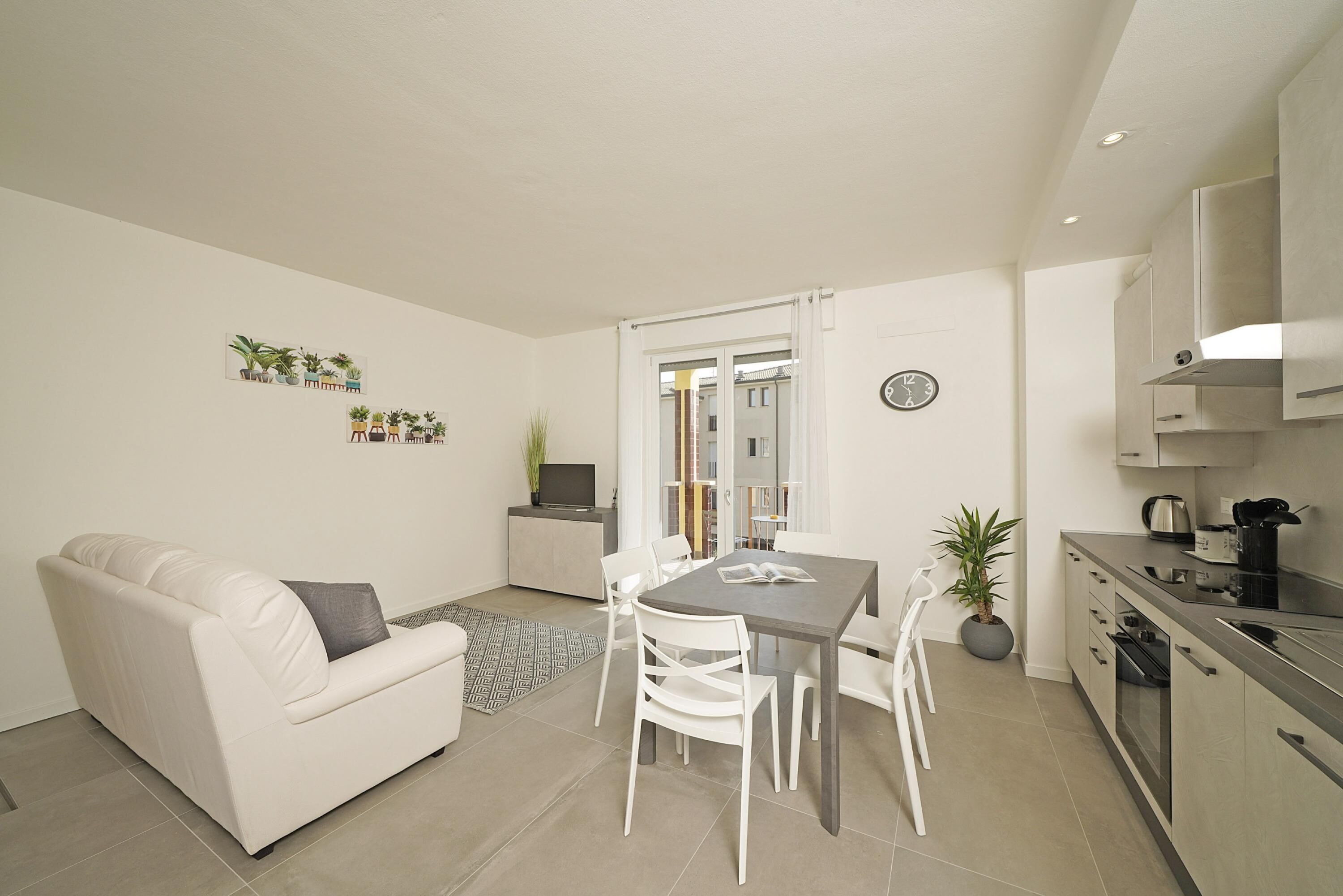A modern living room with a white sofa, dining table, and chairs, a kitchen with stainless steel appliances, and a small potted plant.