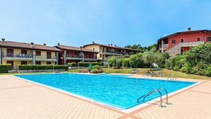 Ein rechteckiger Swimmingpool mit einer gefliesten Sonnenterrasse, umgeben von einem angelegten Garten und mehrstöckigen Gebäuden mit Balkonen.