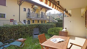 Ein überdachter Außenbereich mit Tisch und Stühlen, eine Treppe, die zu einem Balkon führt, und ein Hecken im Hintergrund.