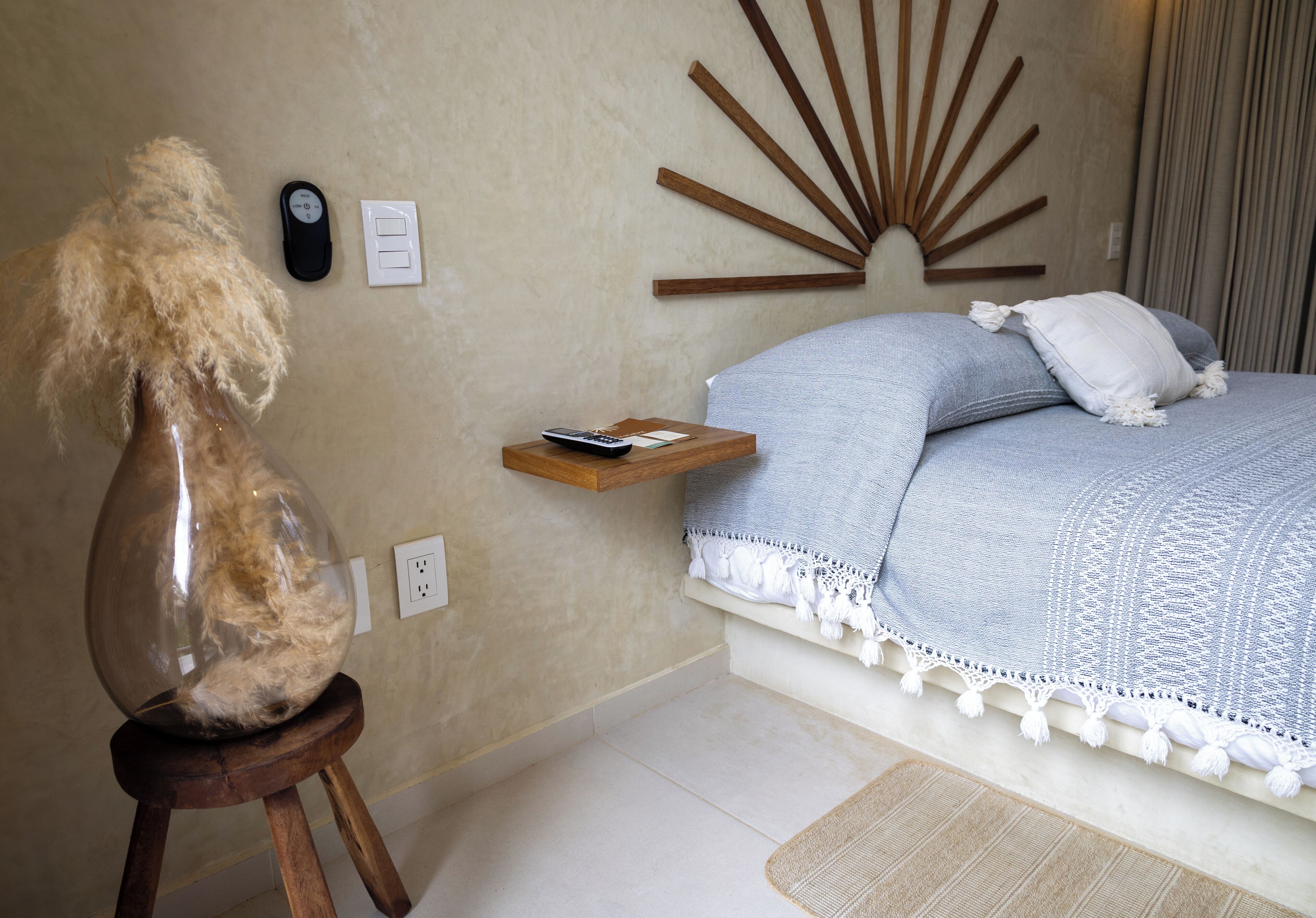 A bedroom with a bed, a bedside table, a wall-mounted light switch, and a decorative wall piece.