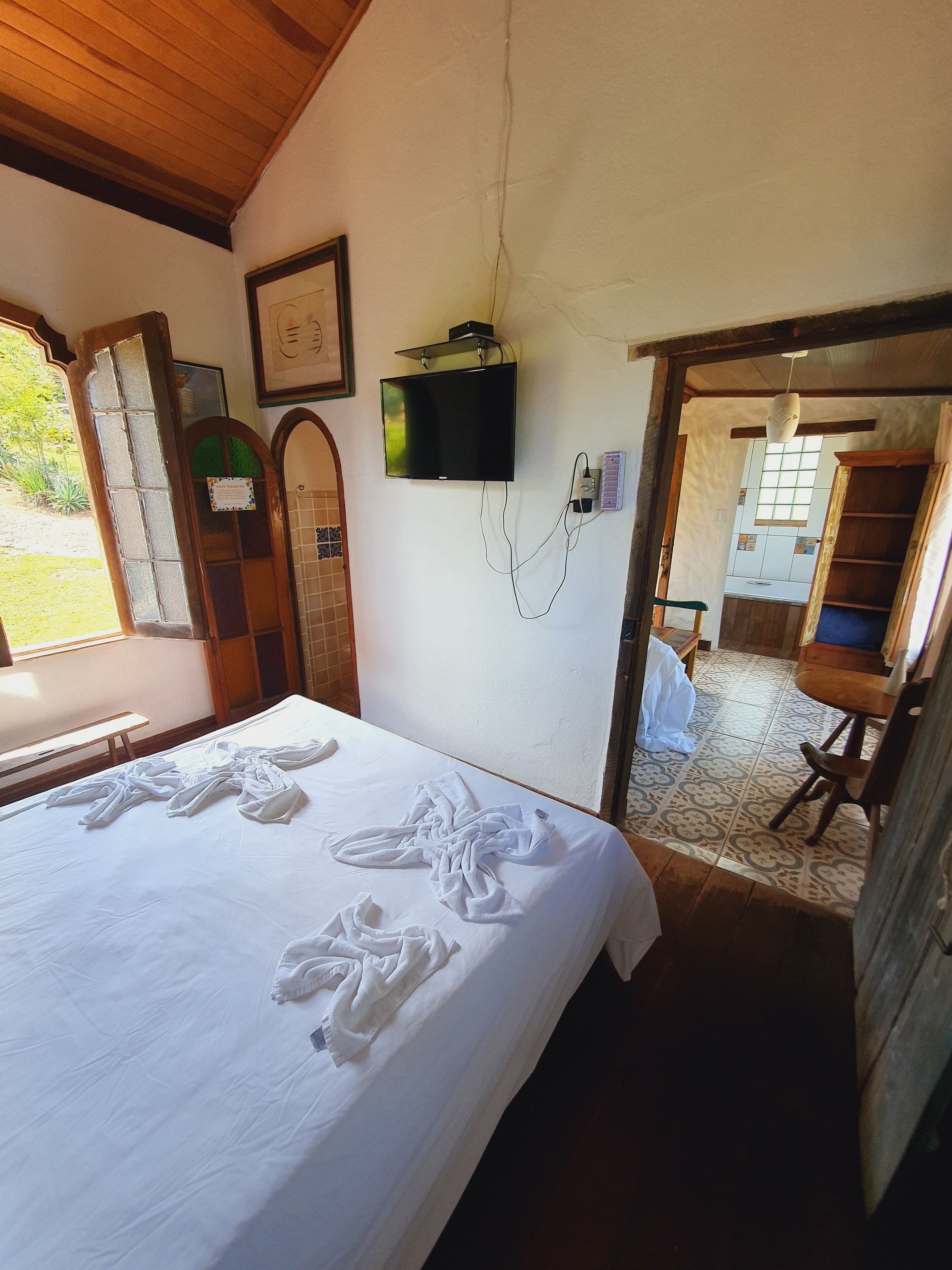 A bedroom with a bed, a television mounted on the wall, and a view of the kitchen through an open doorway.