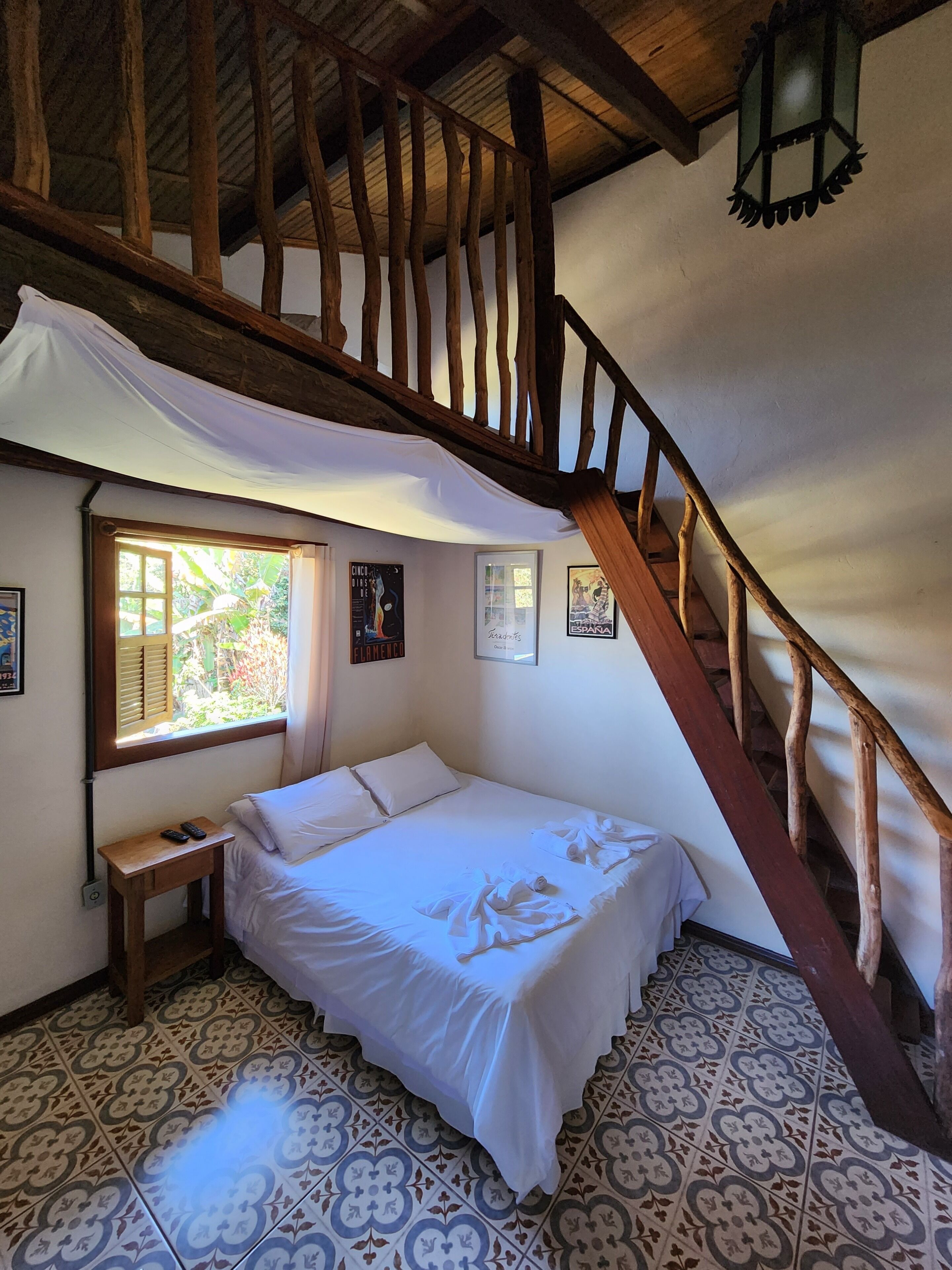 Un dormitorio con cama, una mesita de noche de madera, una ventana con persianas, una escalera y baldosas de suelo con estampado.