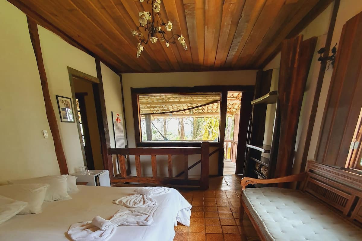 Ein Hotelzimmer mit zwei Betten, einem Sofa, einer Holzbalkendecke und einem Fenster mit Blick auf Grünflächen.