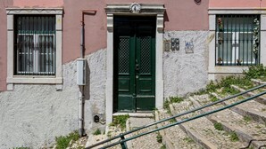 Un bâtiment avec une porte verte, une fenêtre ornée de barreaux en métal et un escalier menant à l’entrée.