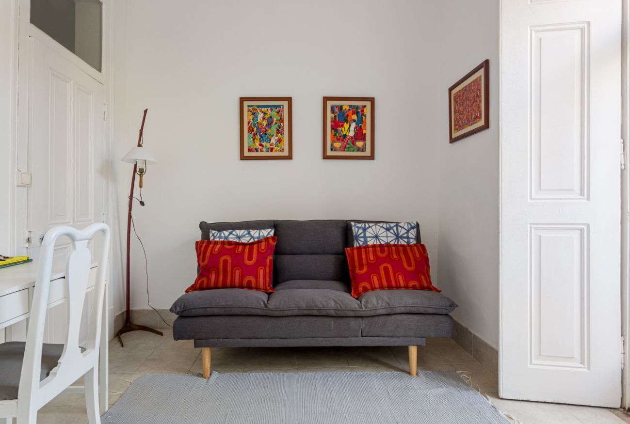 Ein Zimmer mit einem grauen Sofa, zwei farbenfrohen gerahmten Kunstwerken, einer weißen Tür und einem weißen Schreibtisch mit einem Stuhl.