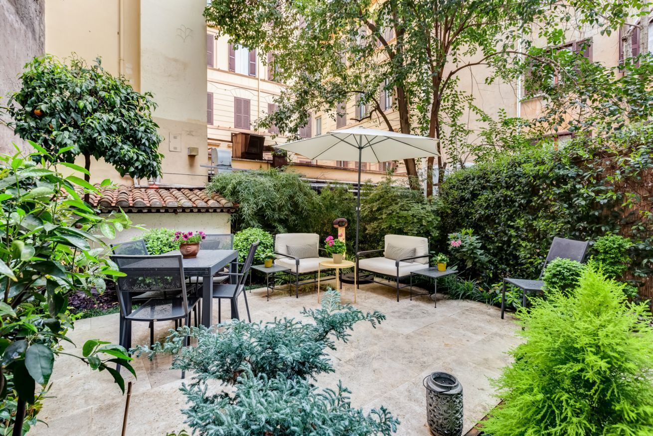 An outdoor patio area with a table and chairs, surrounded by potted plants and a large umbrella.