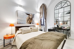 A modern bedroom with a large bed, two bedside lamps, a painting, a desk with a chair, and a large mirror.