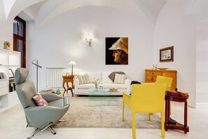 A modern living room with a white sofa, a glass coffee table, a grey armchair, and a yellow chair. There is a wooden cabinet and a small table with a lamp.