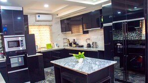 A modern kitchen with dark cabinetry, a built-in microwave, a refrigerator, and a marble countertop. - 5 bed Home w/ Professional Chef, Strong Security, Daily Housekeeping (Abuja)