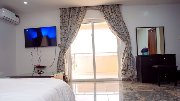 A hotel room with a bed, a flat-screen TV mounted on the wall, a dresser, and a sliding glass door leading to a balcony. - 5 bed Home w/ Professional Chef, Strong Security, Daily Housekeeping (Abuja)